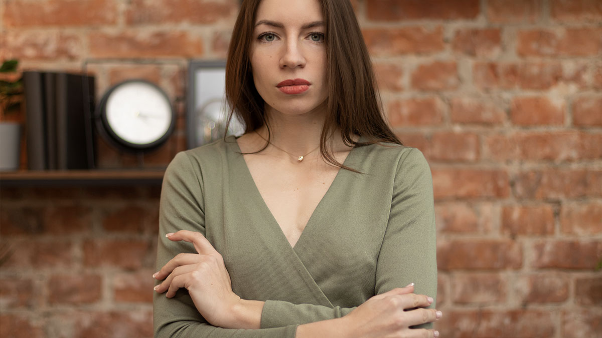 Woman stands her ground with arms crossed, serious expression, facing camera against a brick wall background.