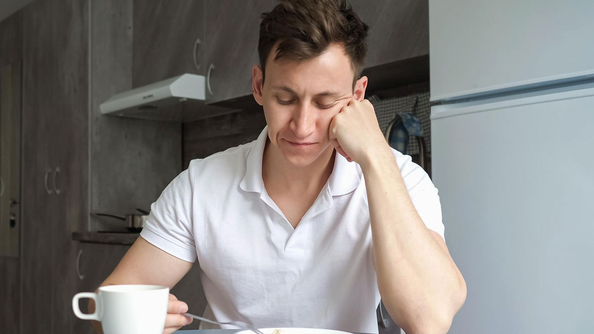 Young man in white shirt looking uninterested while sitting at kitchen table with vegan food and cup.