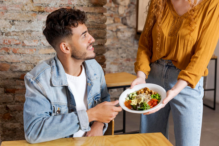 Man hesitating to eat vegan meal offered by girlfriend during birthday celebration at cozy restaurant table.