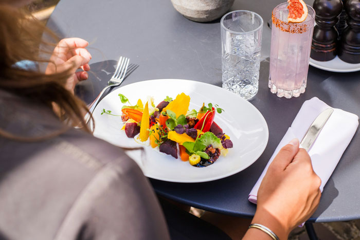 Person sitting at a table with a colorful vegan meal, holding utensils and a glass of water nearby.