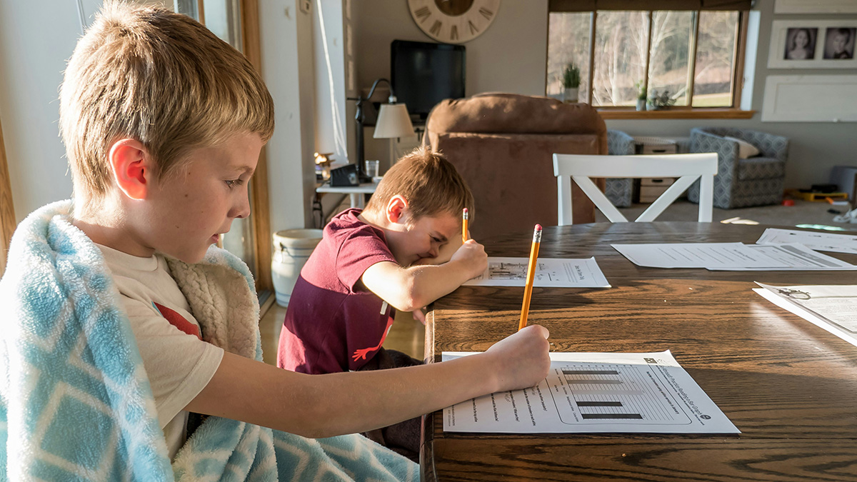 Two boys doing homeschool worksheets at a table in a cozy living room with natural sunlight.