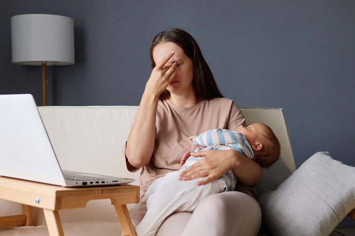 Woman sitting on a couch holding a baby, looking stressed, with a laptop nearby, symbolizing emotions around refusing let rehome cat. Woman sitting on a couch holding a baby, looking stressed, with a laptop nearby, symbolizing emotions around refusing let rehome cat.