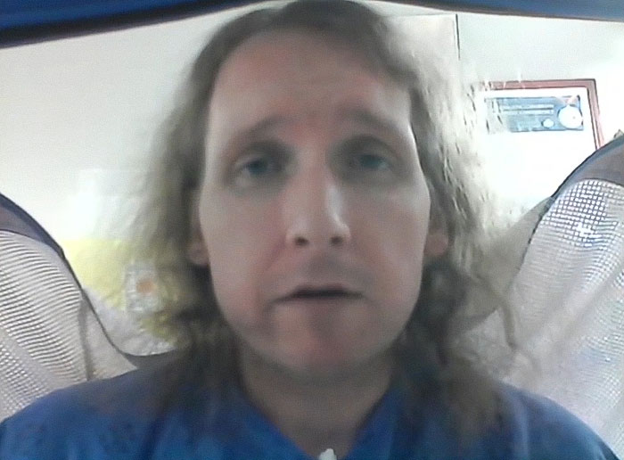 Man with long hair and blue shirt speaking on video call after being trapped in hospital for nine years exposing staff abuse. Man with long hair and blue shirt speaking on video call after being trapped in hospital for nine years exposing staff abuse.