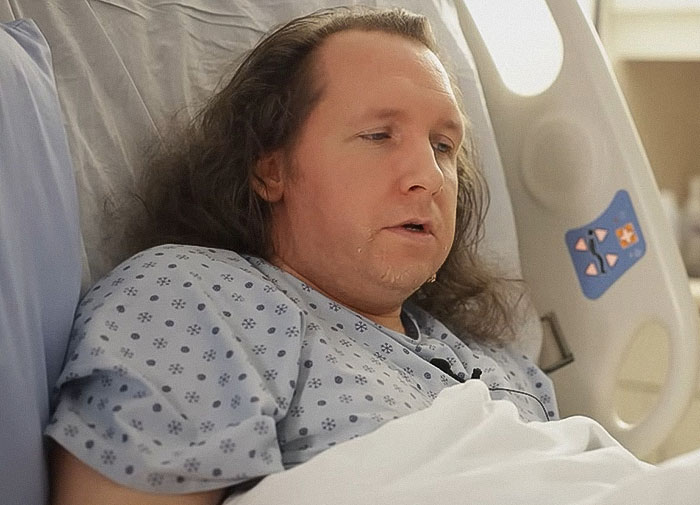 Man lying in hospital bed wearing patterned hospital gown, appearing distressed and fatigued during recovery. Man lying in hospital bed wearing patterned hospital gown, appearing distressed and fatigued during recovery.