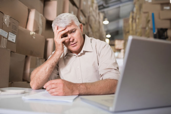 Stressed warehouse manager surrounded by cluttered boxes, holding head in frustration due to backroom disaster after new rule. Stressed warehouse manager surrounded by cluttered boxes, holding head in frustration due to backroom disaster after new rule.
