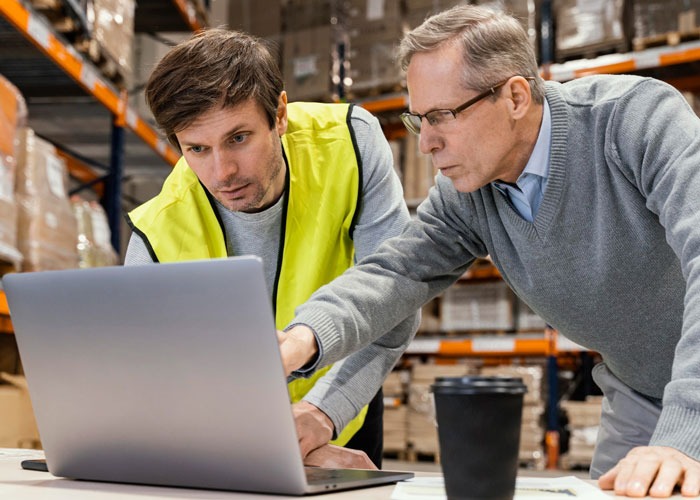 Two men in a warehouse reviewing store management on a laptop, highlighting issues caused by a new manager's rule. Two men in a warehouse reviewing store management on a laptop, highlighting issues caused by a new manager's rule.