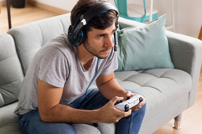 Young man with headset focused on exploratory testing while playing a game and reporting bugs on a gray couch.