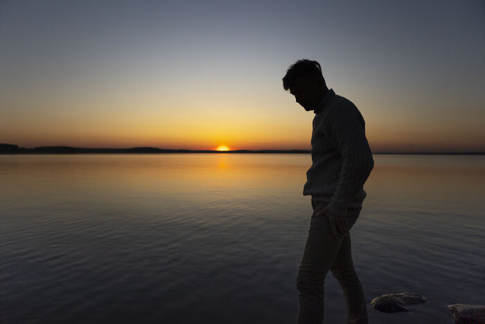 Silhouette of a man reflecting by calm water at sunset, conveying raw emotional confessions and heavy secrets.