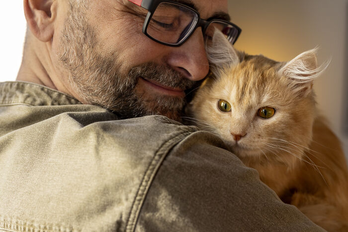 Man with glasses hugging an orange cat, conveying raw and emotional confessions of carrying heavy secrets for years.