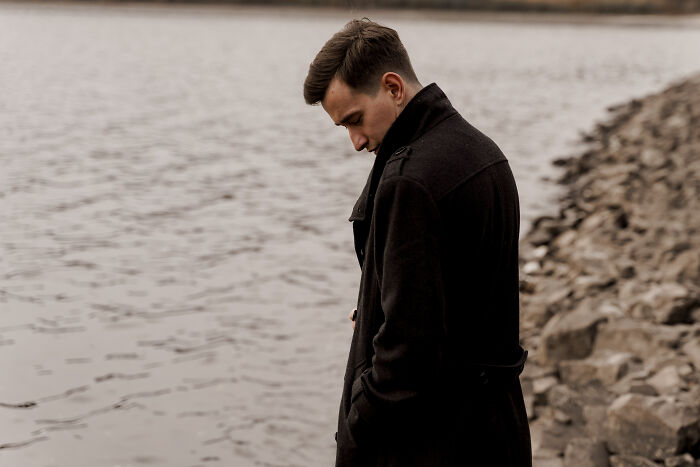 Young man in a dark coat standing by the water, reflecting with raw and emotional confessions about heavy secrets.