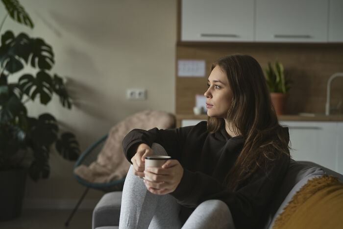 A woman sitting on a couch holding a cup, reflecting deeply in a cozy living room, carrying heavy secrets.