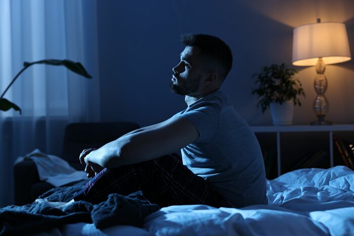 Man sitting on bed at night, appearing deep in thought and carrying heavy secrets in an emotional moment.