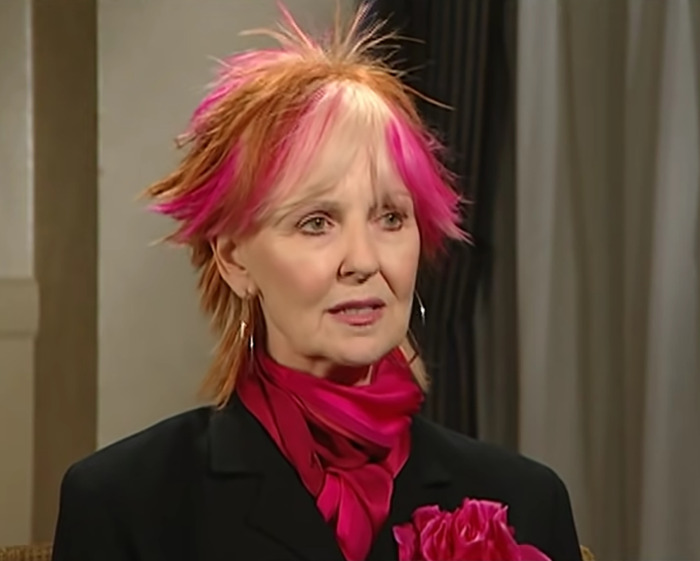 Sitcom legend with vibrant pink and orange hair, wearing black suit and matching pink scarf and flower during interview. Sitcom legend with vibrant pink and orange hair, wearing black suit and matching pink scarf and flower during interview.