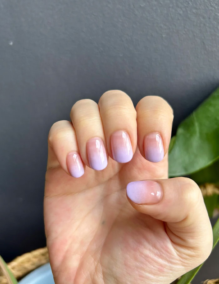 Short French tip nails with a soft purple ombre design on natural nails against a dark gray background and green leaf. Short French tip nails with a soft purple ombre design on natural nails against a dark gray background and green leaf.