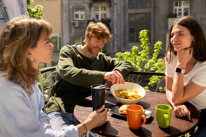 Three friends sitting outdoors at a round table, showing shy body language during casual social interaction.