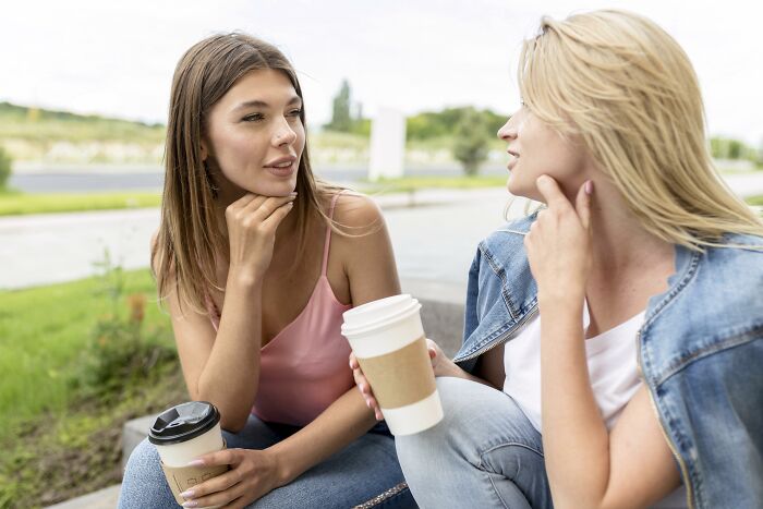Two women sitting outdoors with takeaway coffee cups, engaging in a deep conversation about being shy challenges.