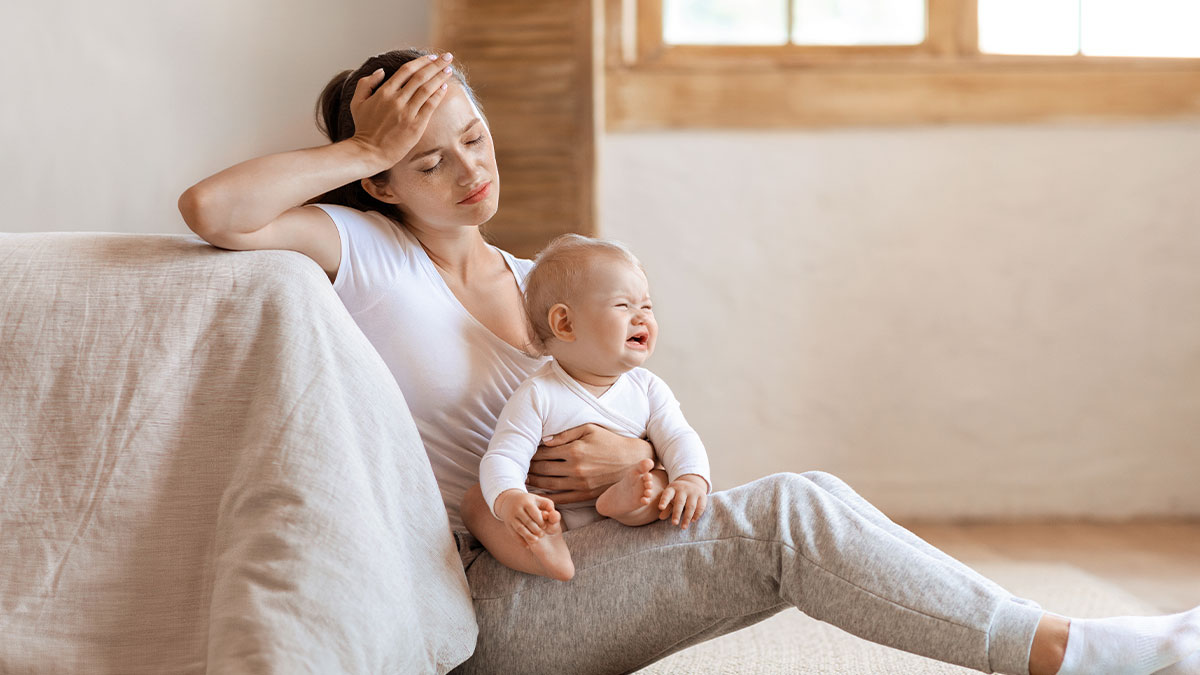 Tired young woman sitting on floor holding crying baby, showing stress of expected babysitting during family visit.