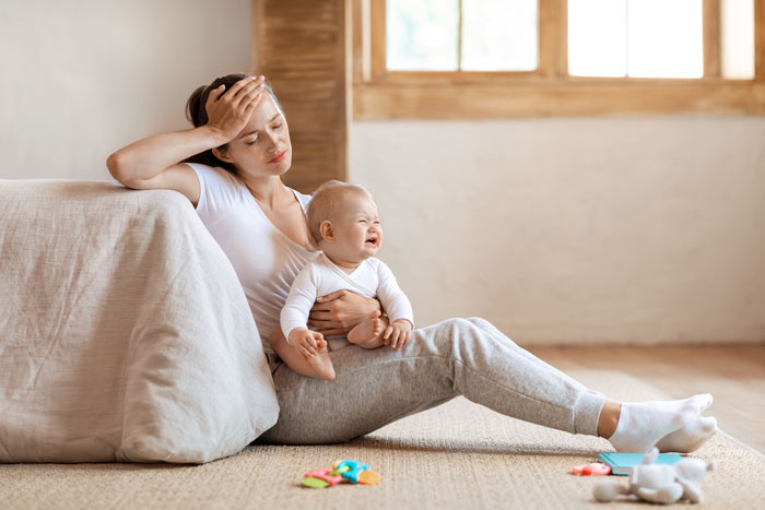 Woman looking tired while sitting on the floor holding a crying baby, highlighting babysitting challenges during family visit. Woman looking tired while sitting on the floor holding a crying baby, highlighting babysitting challenges during family visit.