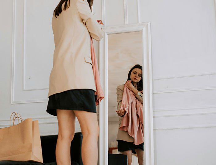 Young woman holding a pink dress in front of a mirror, looking thoughtful in a room before a birthday dinner sabotage. Young woman holding a pink dress in front of a mirror, looking thoughtful in a room before a birthday dinner sabotage.