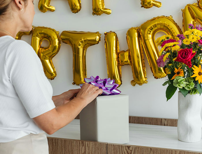 Woman wrapping birthday gift with purple bow near flowers and gold birthday balloons, hinting at sister birthday dinner sabotage Woman wrapping birthday gift with purple bow near flowers and gold birthday balloons, hinting at sister birthday dinner sabotage
