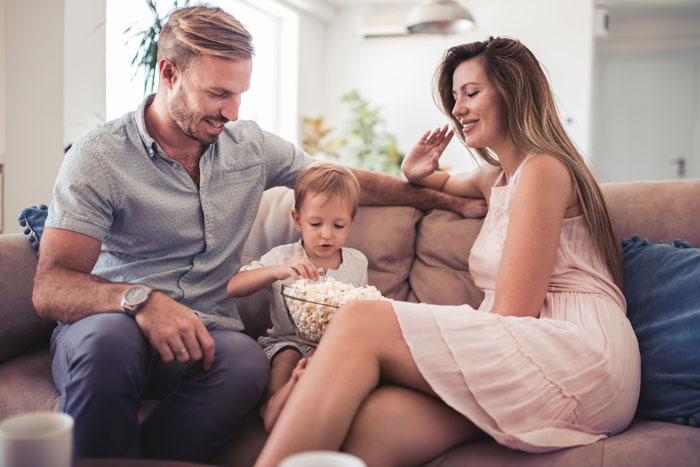Child-free lady warning sister about too much babysitting while sitting on couch with man and child sharing popcorn. Child-free lady warning sister about too much babysitting while sitting on couch with man and child sharing popcorn.