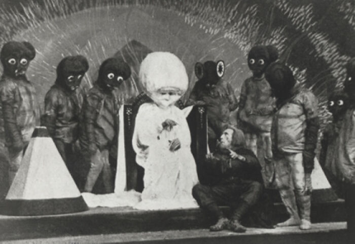 Black and white eerie old photo showing a group in creepy costumes with large eyes surrounding a seated figure in white, unsettling past scene.