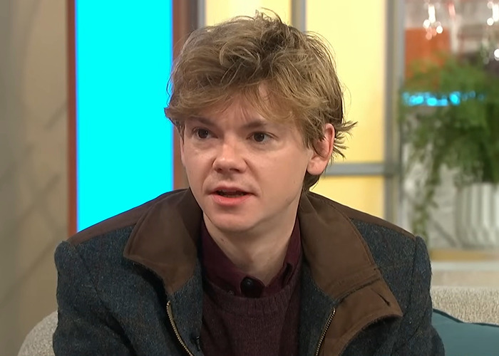Young man with tousled hair wearing a dark coat during a daytime TV interview discussing viral photos of Sean Connery. Young man with tousled hair wearing a dark coat during a daytime TV interview discussing viral photos of Sean Connery.
