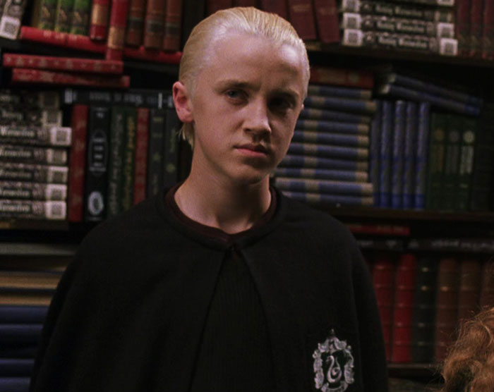 Tom Felton as Draco Malfoy in Slytherin robes standing in front of bookshelves in a Harry Potter scene. Tom Felton as Draco Malfoy in Slytherin robes standing in front of bookshelves in a Harry Potter scene.