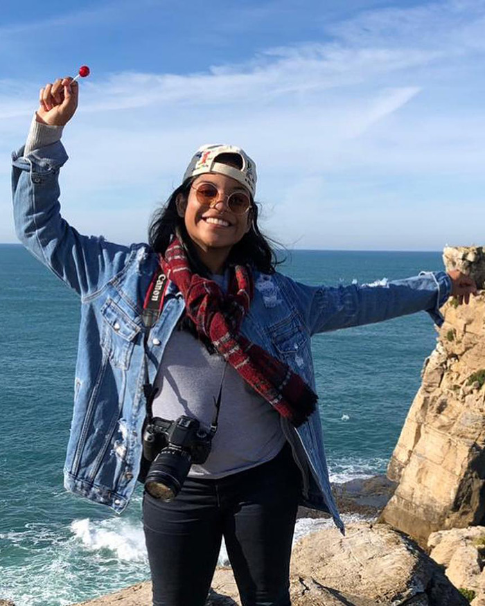Smiling tourist wearing sunglasses and denim jacket, standing on rocky cliff by ocean, with camera around neck and candy in hand. Smiling tourist wearing sunglasses and denim jacket, standing on rocky cliff by ocean, with camera around neck and candy in hand.