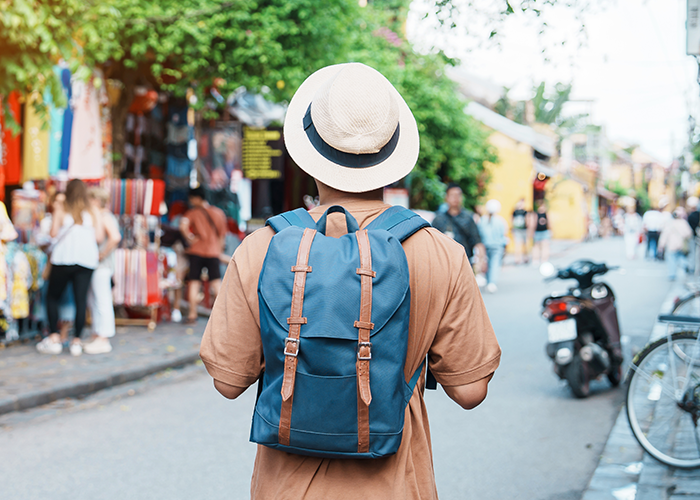 Tourist with backpack and hat walking through busy street near historic site with 2000-year-old statues.