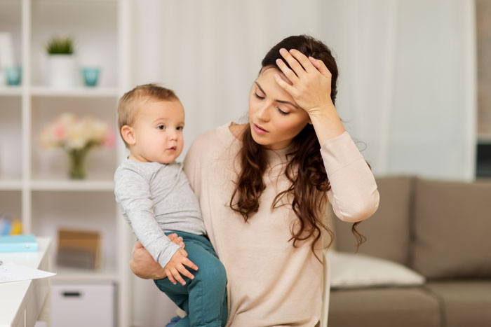 Tired mother holding young child indoors, illustrating the challenges of vacation without kids babysitting options.