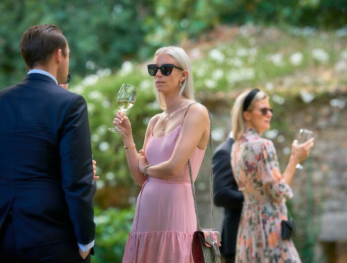 Group of elegantly dressed middle class people socializing outdoors, holding drinks in a relaxed garden setting.