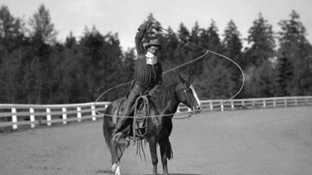 Vintage cowboy on horseback skillfully twirling a lasso in an outdoor rodeo arena with trees in the background