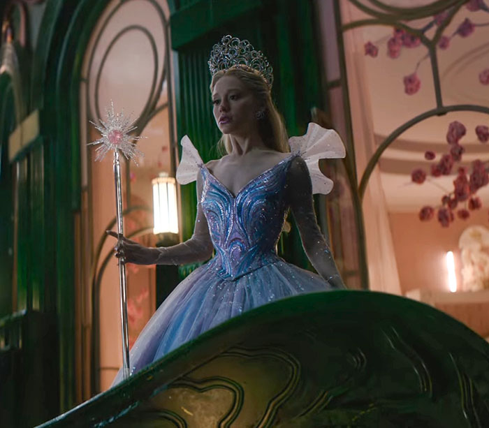 Ariana Grande in a blue gown and tiara holding a wand, showcasing an entirely visible ribcage in a cinematic scene. Ariana Grande in a blue gown and tiara holding a wand, showcasing an entirely visible ribcage in a cinematic scene.