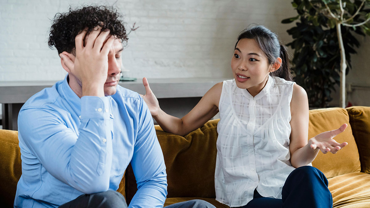 Wife explaining herself emotionally to husband sitting upset on couch, referencing mind control as an excuse.