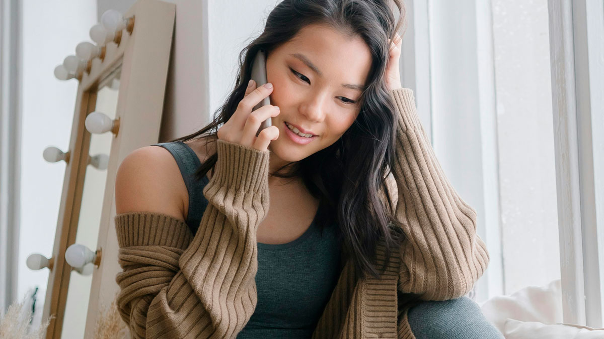 Young woman in cozy sweater talking on phone by window, contemplating refusing wifeu2019s work husband on family vacation