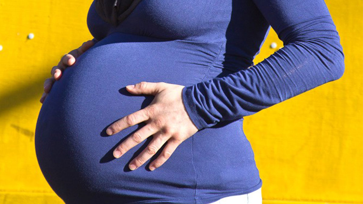 Pregnant woman in a blue shirt holding her twin pregnancy belly against a bright yellow background.