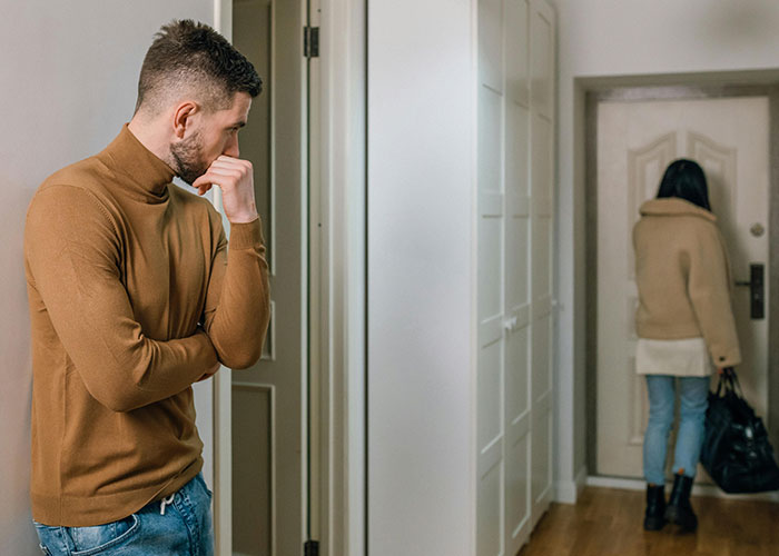 Man in brown sweater looking thoughtful while woman walks away carrying a bag, reflecting a tradwife relationship conflict. Man in brown sweater looking thoughtful while woman walks away carrying a bag, reflecting a tradwife relationship conflict.