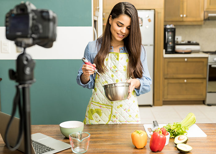 Woman in apron preparing food in kitchen for tradwife lifestyle video recording with camera and laptop nearby Woman in apron preparing food in kitchen for tradwife lifestyle video recording with camera and laptop nearby