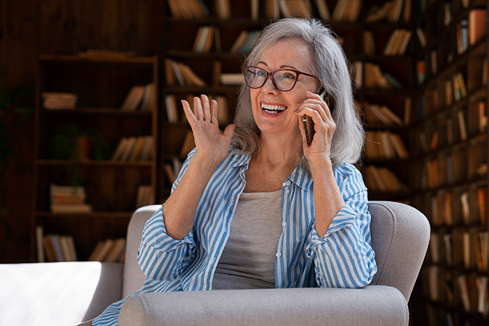 Smiling woman move Alaska alone talking on phone, wearing glasses and striped shirt, sitting in cozy room with bookshelves. Smiling woman move Alaska alone talking on phone, wearing glasses and striped shirt, sitting in cozy room with bookshelves.