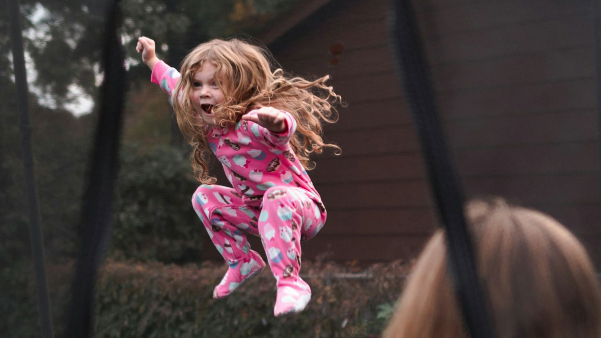 Young girl joyfully jumping on trampoline while woman watches, highlighting family conflict over broken leg claim.