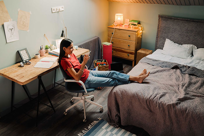 Young woman in casual clothes relaxing in bedroom, focused on her phone, with cozy lighting and a study desk nearby. Young woman in casual clothes relaxing in bedroom, focused on her phone, with cozy lighting and a study desk nearby.