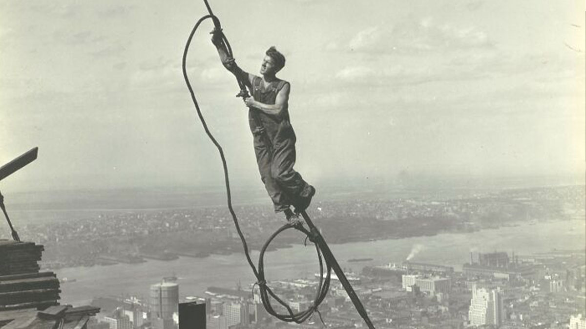 American working class man in vintage photo balancing on high steel beam during construction job above cityscape.