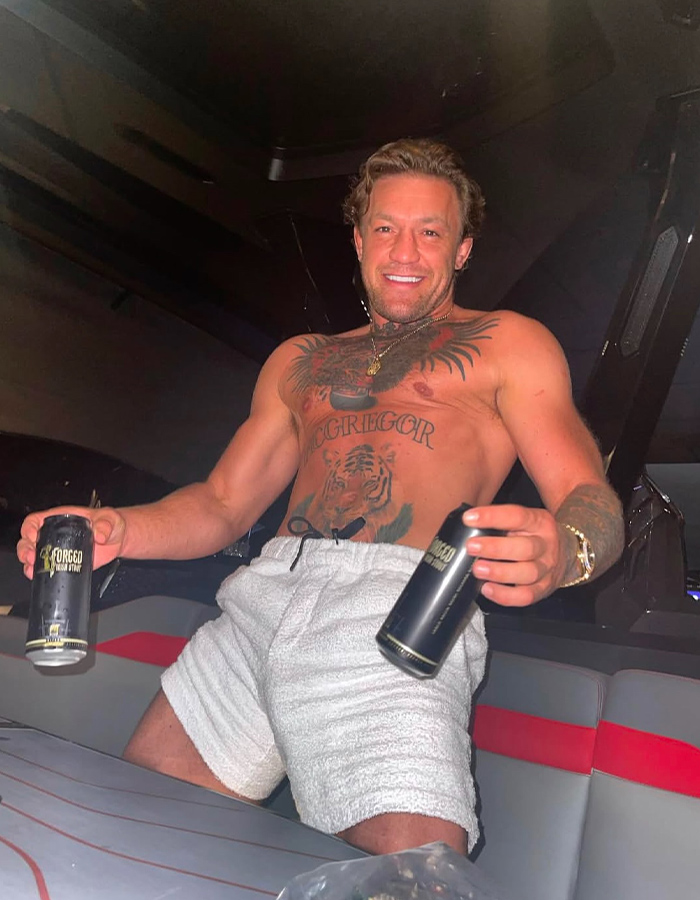 Shirtless man with tattoos smiling and holding two beverage cans in a dimly lit indoor setting. Shirtless man with tattoos smiling and holding two beverage cans in a dimly lit indoor setting.