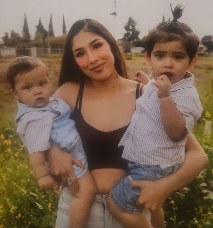 Young mother holding two toddlers outdoors, related to after baby lost his life in car incident and court charges.