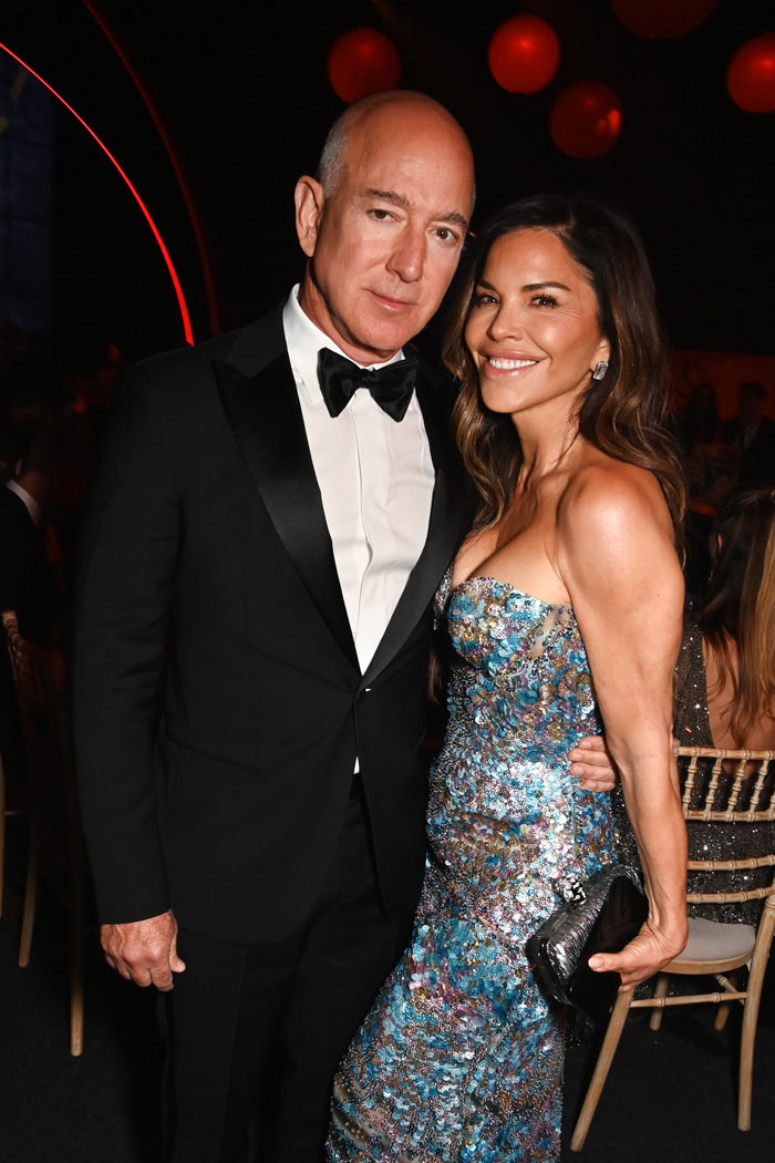 Jeff Bezos wearing a tuxedo with hair, posing with Lauren Sánchez in a colorful dress at an evening event. Jeff Bezos wearing a tuxedo with hair, posing with Lauren Sánchez in a colorful dress at an evening event.
