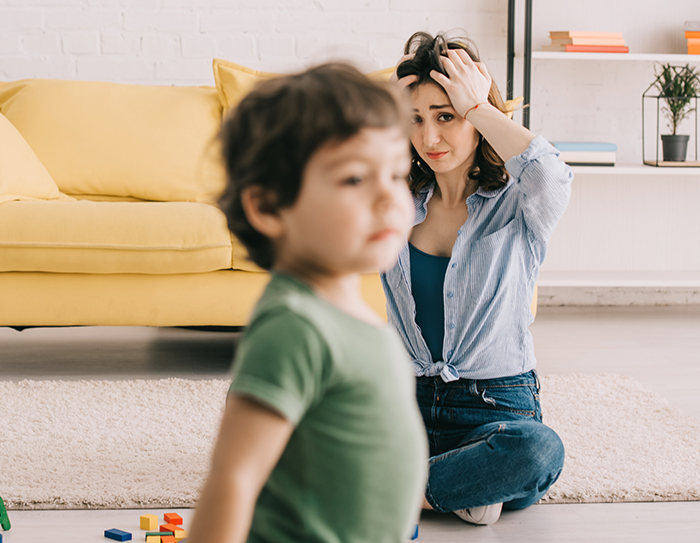 Woman sitting on floor stressed while babysitting toddler in living room with yellow couch in background Woman sitting on floor stressed while babysitting toddler in living room with yellow couch in background