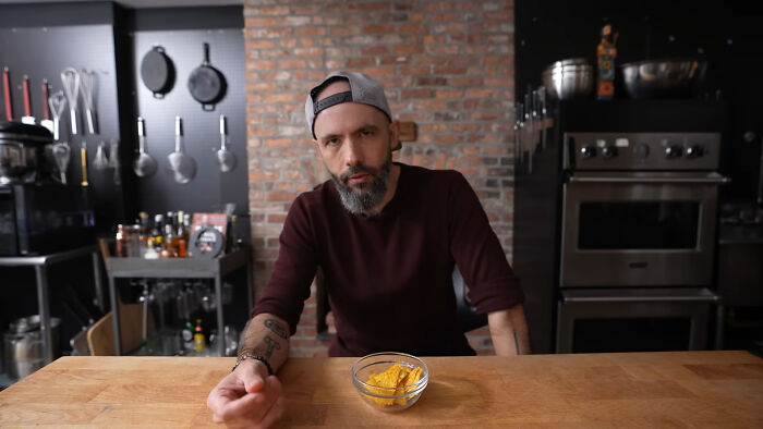 Man wearing a backward cap sitting at kitchen table with bowl of chips, discussing people share unsubscribed YouTubers.
