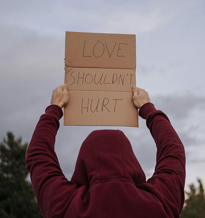 Person in a maroon hoodie holding a sign with love shouldn't hurt message, symbolizing domestic violence awareness and justice. Person in a maroon hoodie holding a sign with love shouldn't hurt message, symbolizing domestic violence awareness and justice.