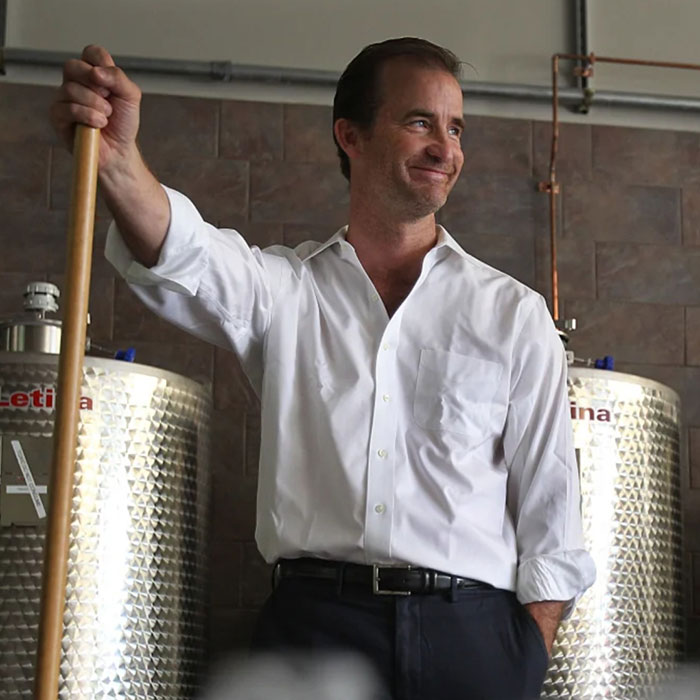 Man in white shirt leaning on a wooden stick, smiling inside an industrial setting with metal tanks in the background. Man in white shirt leaning on a wooden stick, smiling inside an industrial setting with metal tanks in the background.